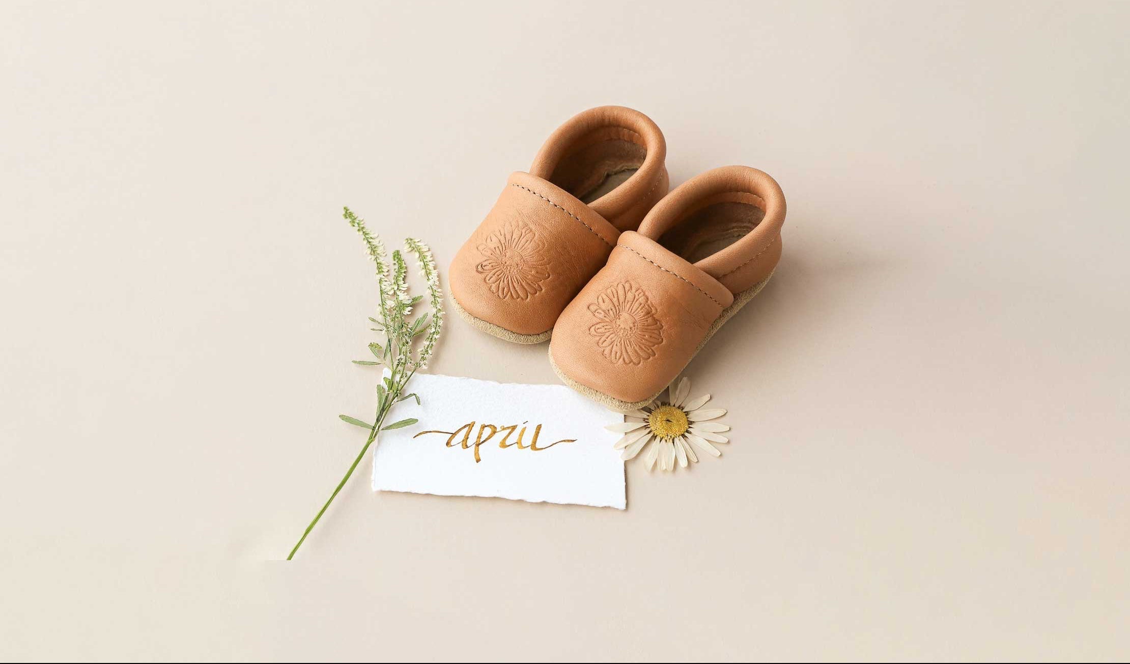 Pair of brown slippers on a light beige background with a small card and April birth flower