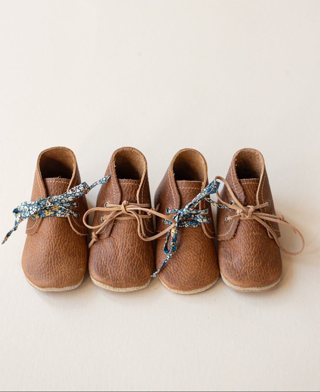 Four small brown leather shoes arranged in a row on a plain light beige background, each tied with a decorative blue and gold patterned ribbon.