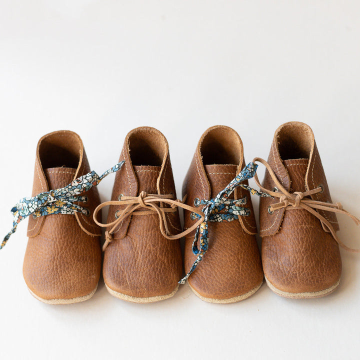 Four tan leather baby boots arranged in a row on a white background, each tied with a decorative blue patterned ribbon.