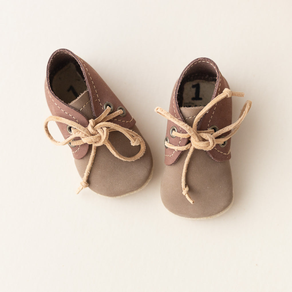 A pair of  baby shoes  oxfords in taupe and brown suede with beige laces, displayed on a clean off-white background.