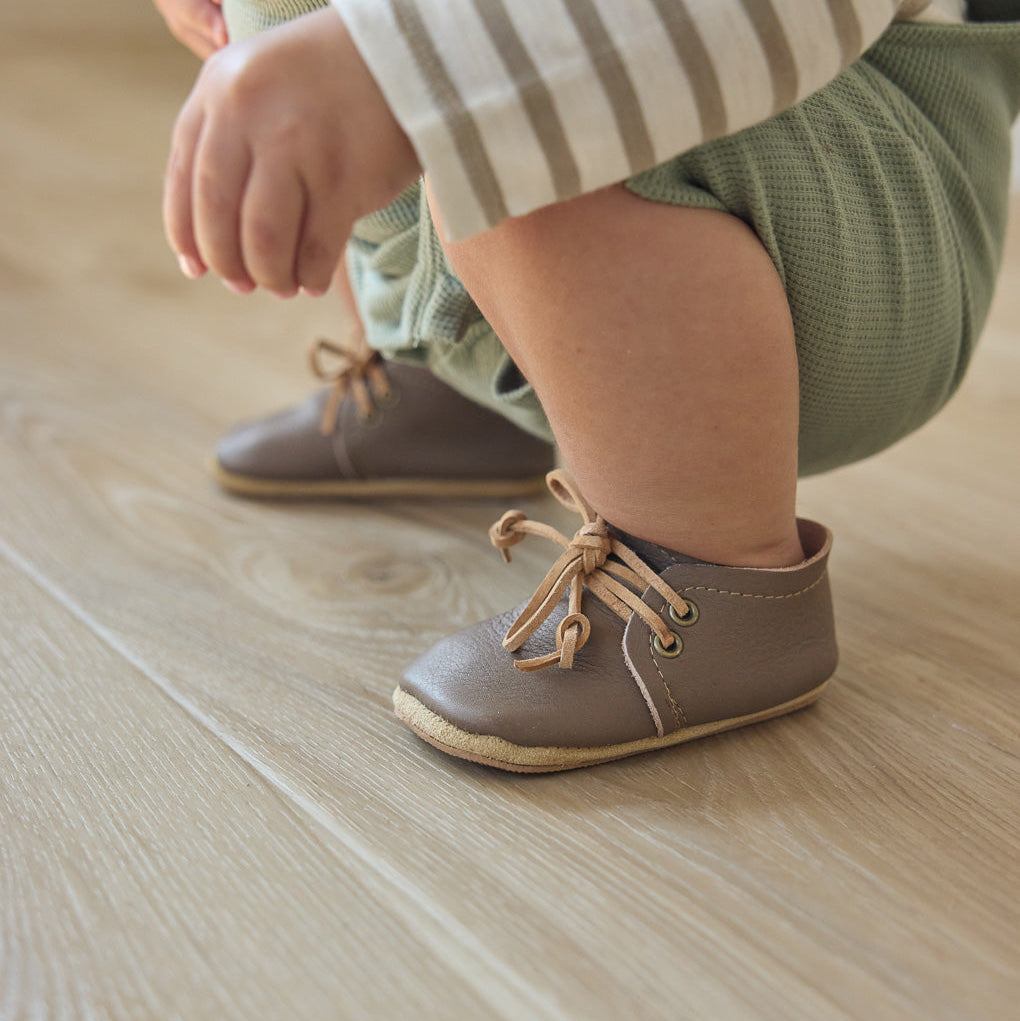 lifetyle image showing taupe baby oxfords on toddler feet. wood floor. 