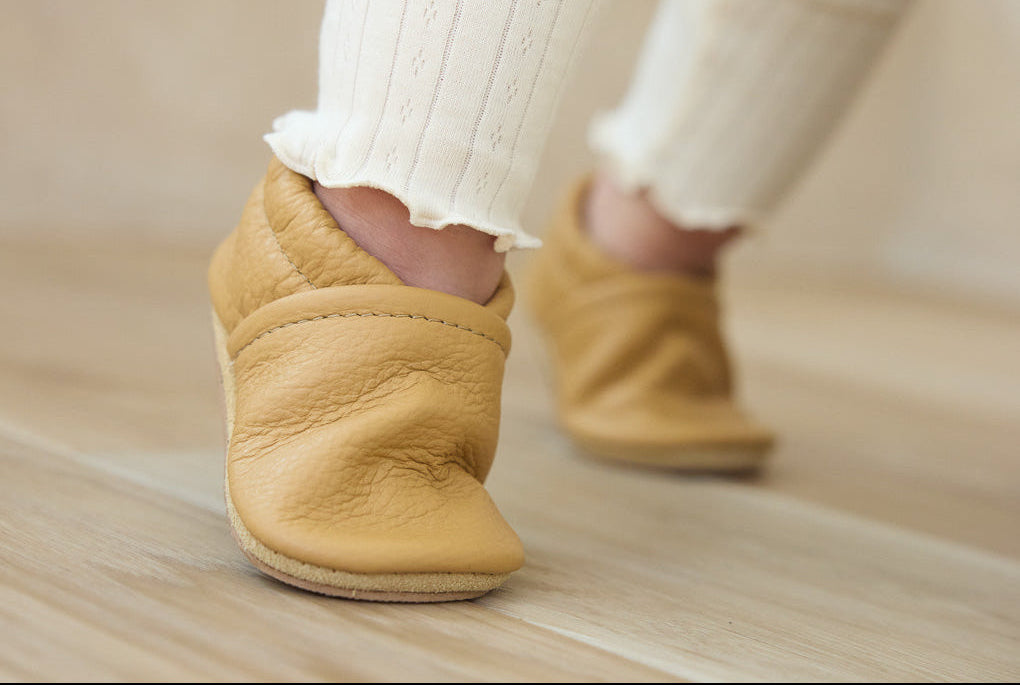 A close-up of a child's feet wearing cream ribbed knit leg warmers and mustard yellow leather baby booties on a light wood floor.