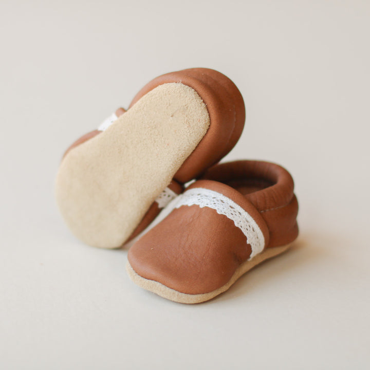product picture of medium brown leather baby girl slip-on shoes with lace trim.  one shoe up to show suede soles.