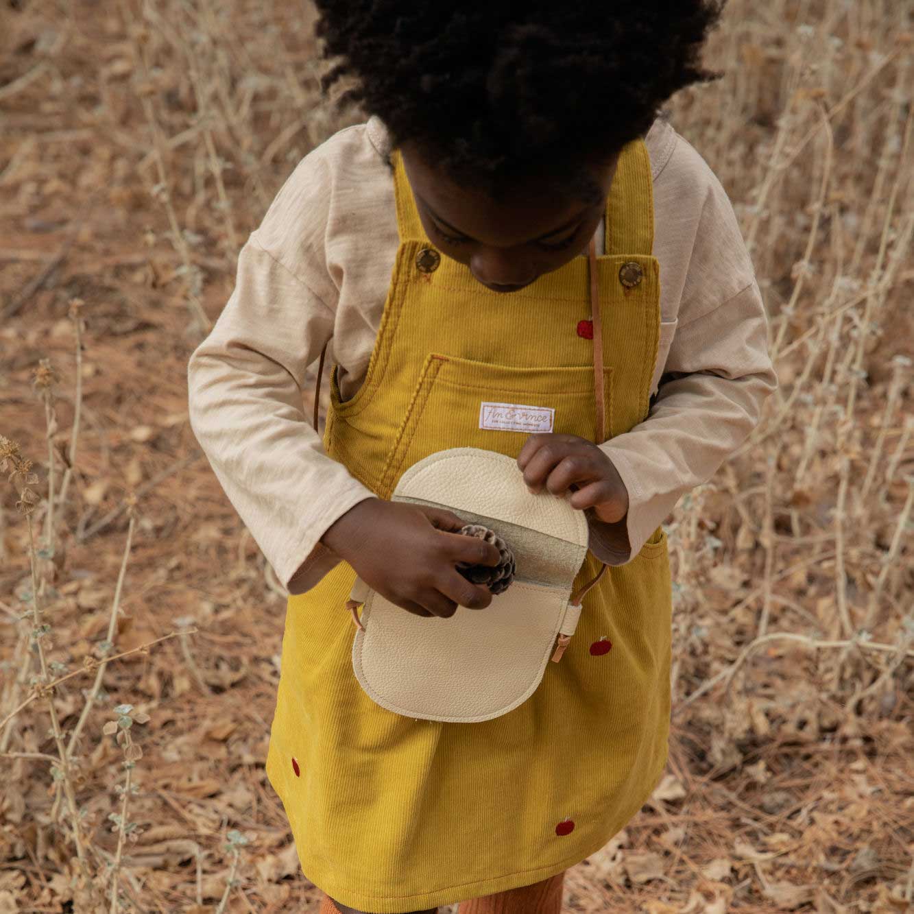 little girl crossbody purse in cream leather. girl putting pine cone inside