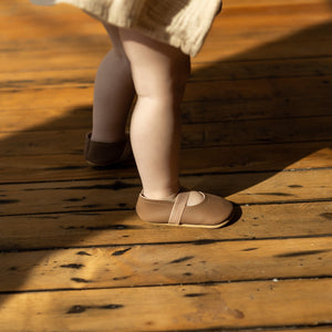 clay and blush leather baby Mary Janes on toddler foot. Distressed wood floor.